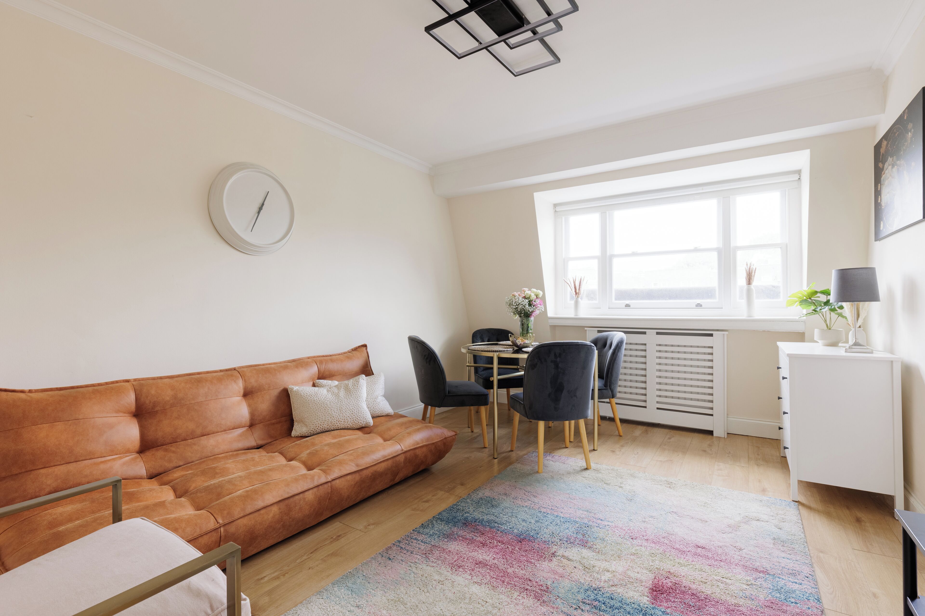 A modern living room with a brown leather sofa, a dining table with chairs, a colorful rug, a white radiator, and a window with natural light.
