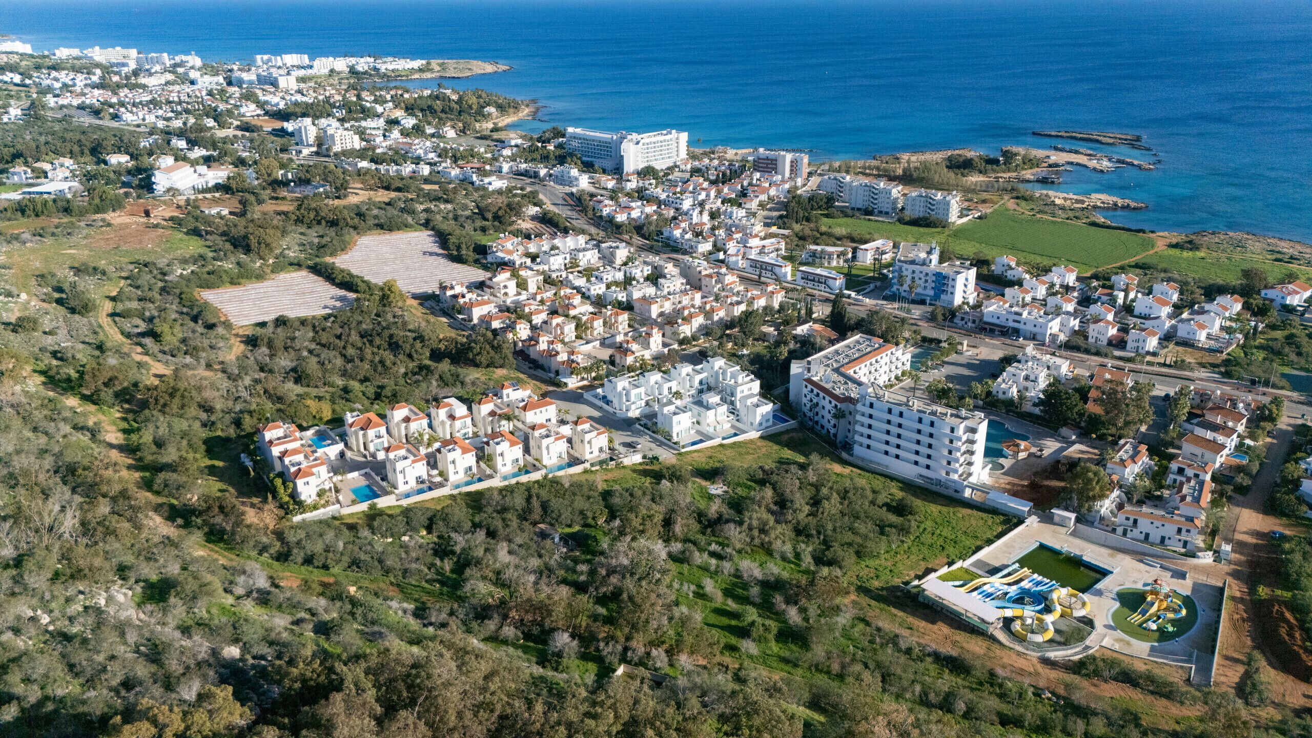 An aerial view of a coastal residential area with white buildings, a large hotel, and a swimming pool complex.