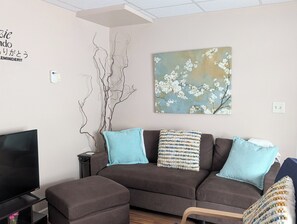 A living room with a brown sofa, two blue cushions, a flat-screen TV, and a wall-mounted painting of white flowers. - Downtown HideAway-  2bed 1st Floor, W/D near UPMC (Williamsport)