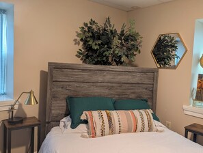 Une chambre avec une tête de lit en bois, un lit avec une literie blanche et un couvre-lit à motifs, une table de chevet avec une lampe et une grande plante en pot.