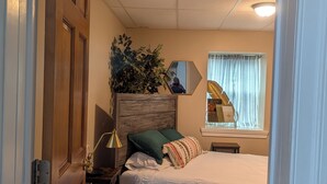 A bedroom with a wooden headboard, a bedside table with a lamp, a green pillow, a striped pillow, and a window with curtains. - Downtown HideAway-  2bed 1st Floor, W/D near UPMC (Williamsport)