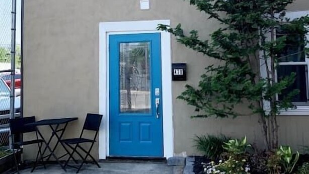 Une porte bleue avec un panneau de verre, un petit espace extérieur aménagé avec une table et des chaises, et un pot de fleurs placé à droite.