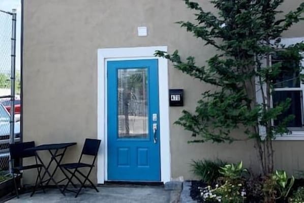 Een blauwe deur met een ruit, een klein terras met een tafel en stoelen, en een bloempot aan de rechterkant.