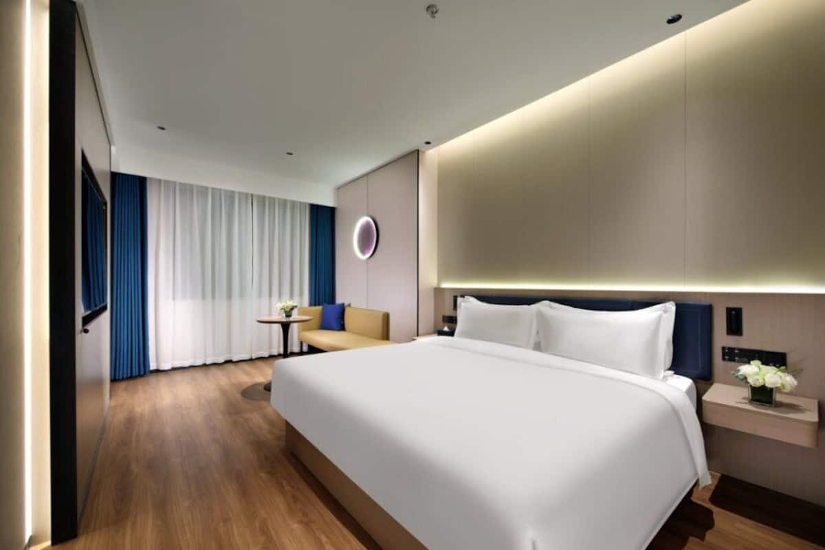 Ein modernes Hotelzimmer mit einem großen Bett, einem Nachttisch mit Blumenvase, einem kleinen runden Tisch mit Stuhl und Blick auf die Stadt durch große Fenster.