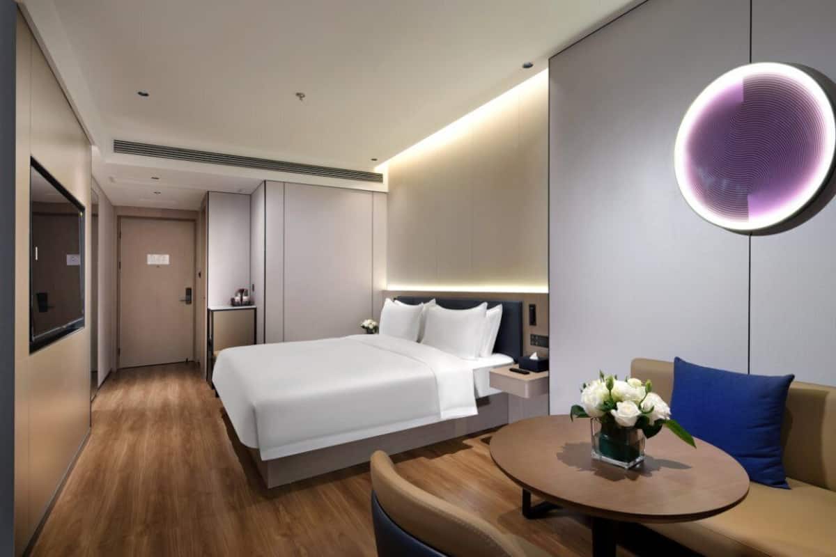 Ein modernes Hotelzimmer mit einem großen Bett, Nachttischen, einem runden Tisch mit Blumenvase und einer Wandleuchte.