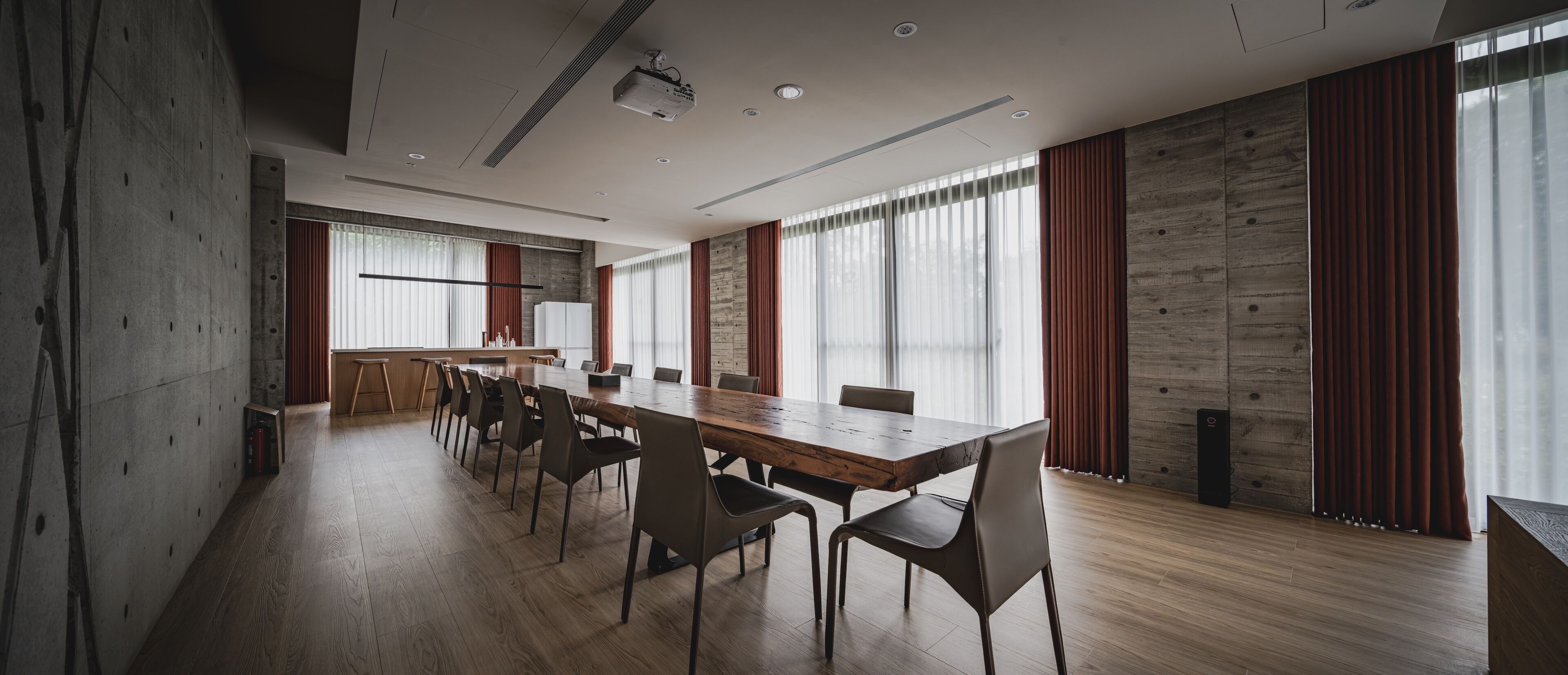 Un salón de conferencias con una larga mesa, sillas, un proyector y ventanas amplias con cortinas.