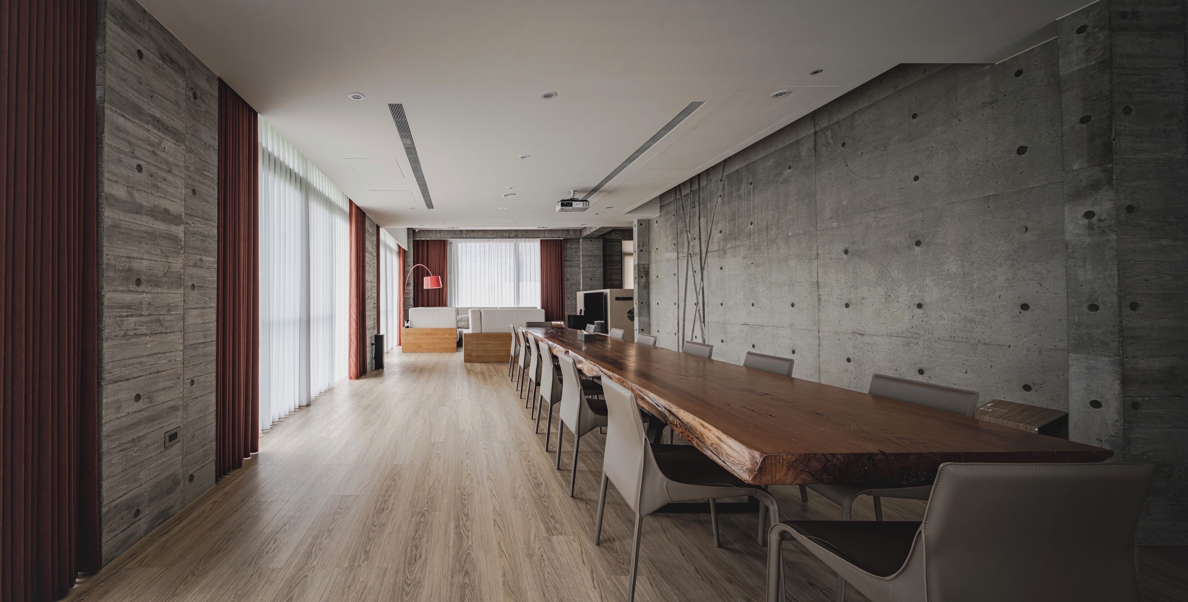 Uma sala de conferências moderna com uma longa mesa de madeira, cadeiras brancas e uma parede de concreto como destaque.