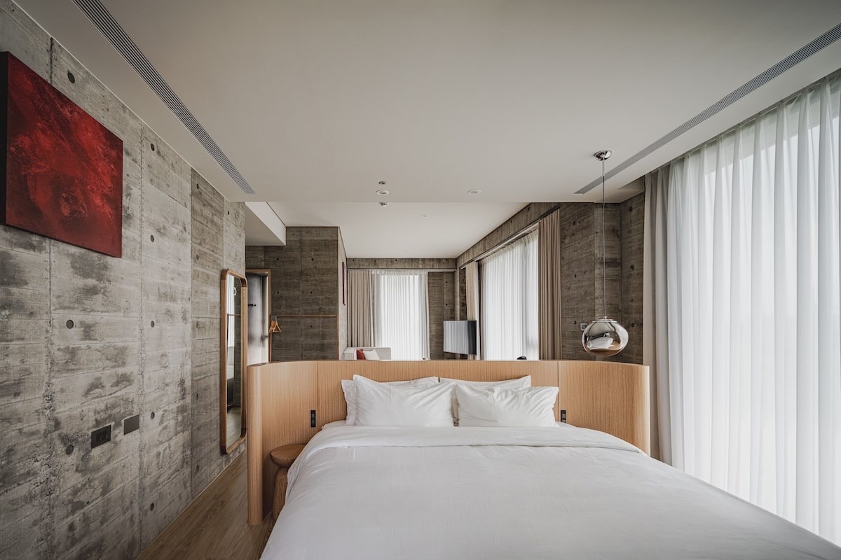 A modern hotel room with a large bed, a wooden headboard, a red abstract painting, and large windows with sheer curtains.