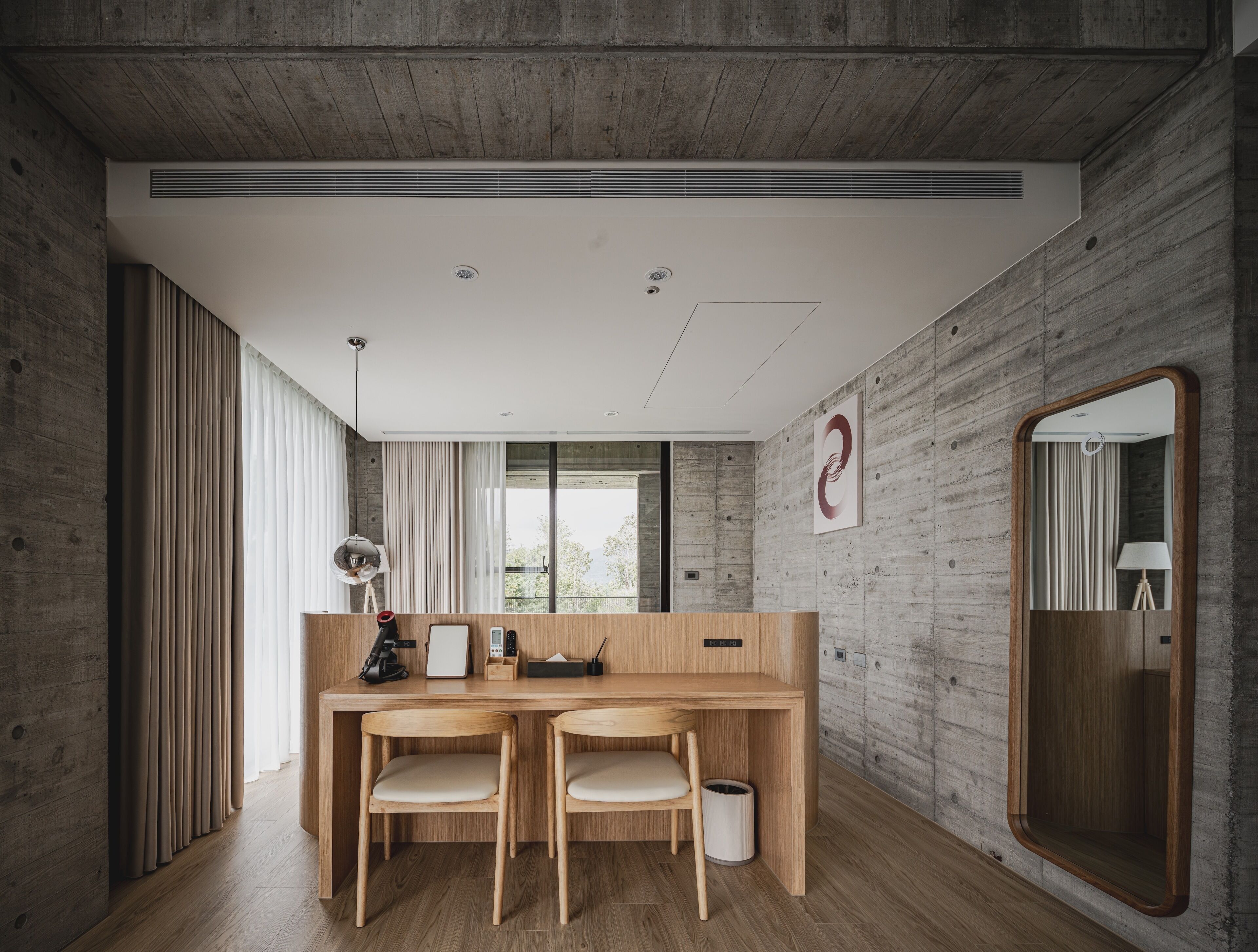 Una habitación moderna con un escritorio de madera, dos sillas, un espejo grande y vistas a árboles por la ventana.