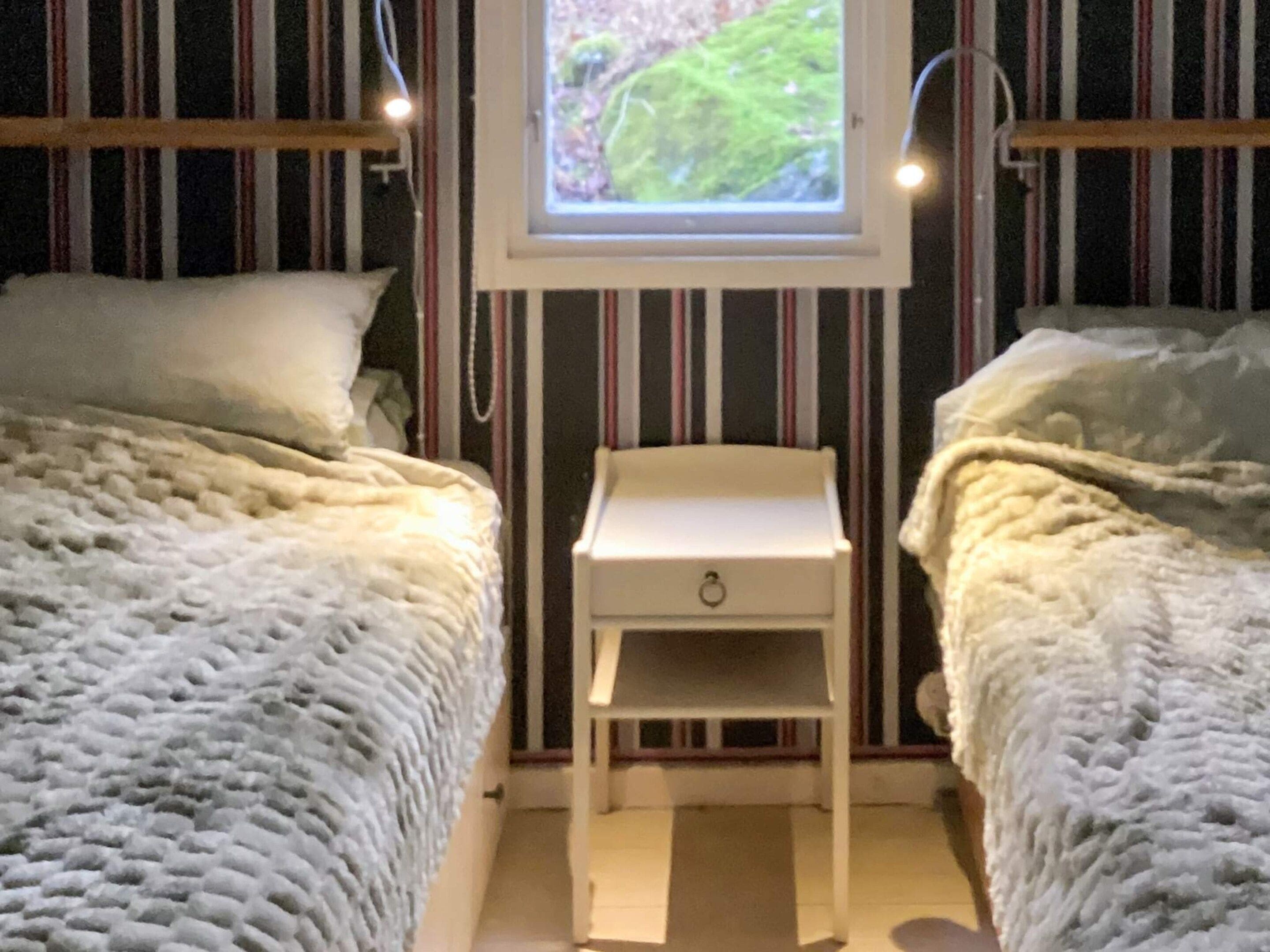 A bedroom with two beds, a small bedside table, and a window with a view of greenery.