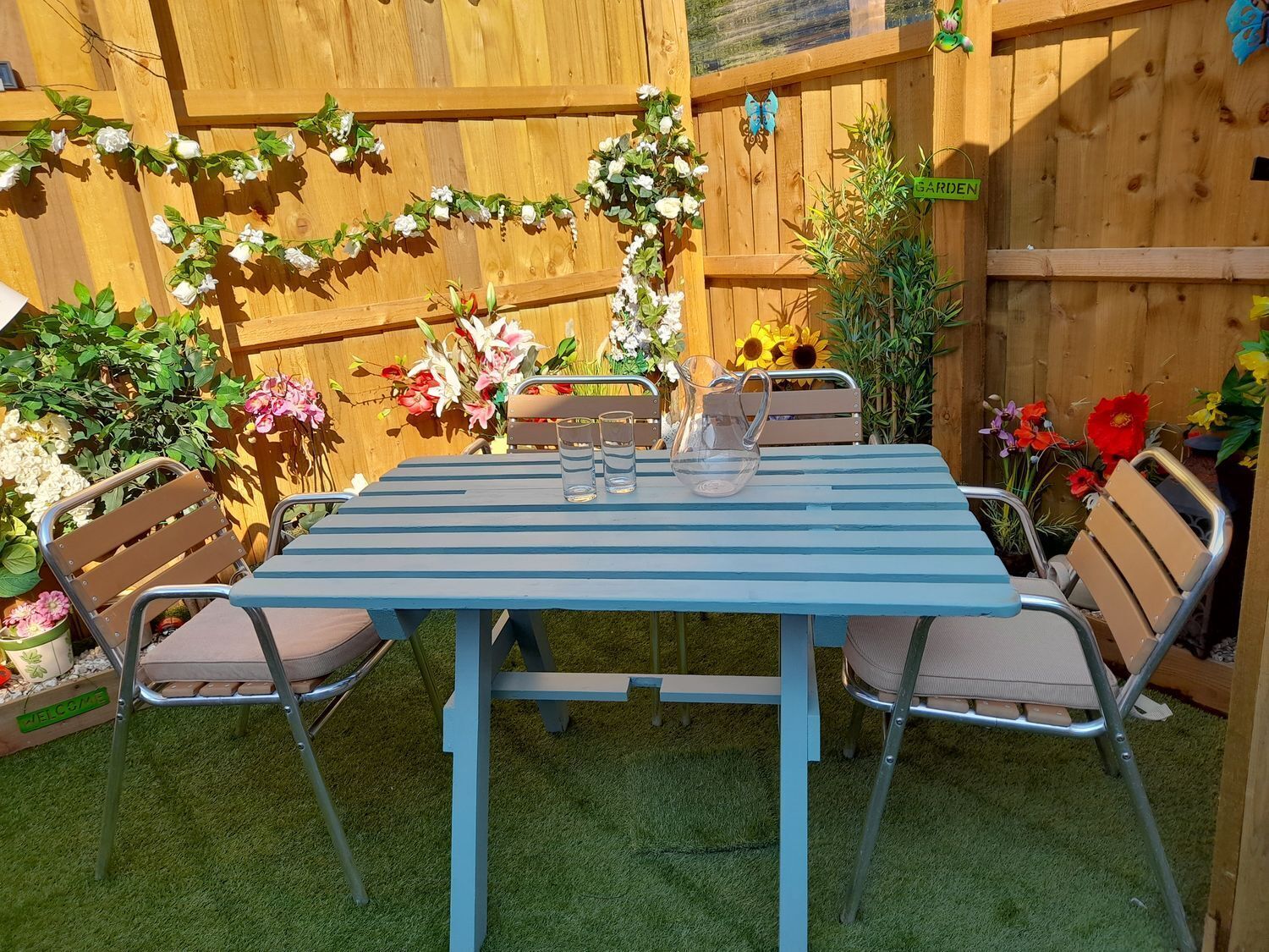 A blue metal table with four chairs, surrounded by a wooden fence with various plants and flowers.