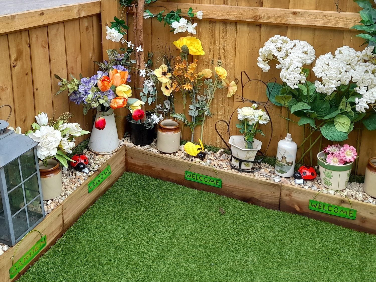 Eine hölzerne Gartenecke mit künstlichen Blumen in Töpfen, einem Willkommensschild und einem kleinen Gartenbereich mit Kunstgrashalm.