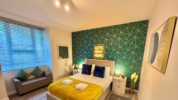 A modern bedroom with a geometric-patterned wall, a bed with a yellow blanket, a sofa with cushions, a bedside table with a lamp, and a 'Let's Stay Home' sign. - Spacious Apartment - With Parking - Town Centre Location - Walk to Beach! (Bournemouth)
