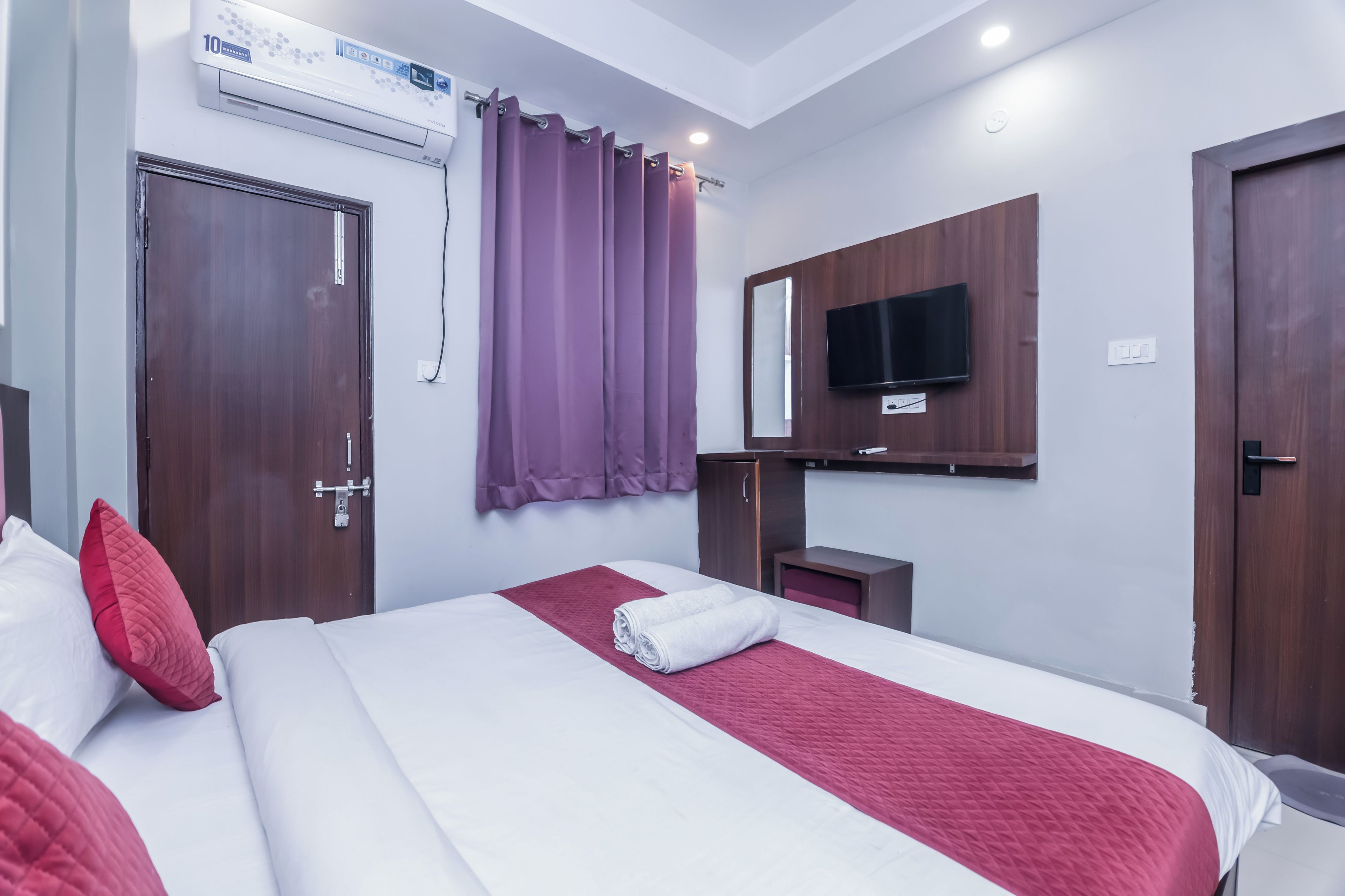 A hotel room with a bed, a TV mounted on the wall, a wardrobe, and a window with purple curtains.