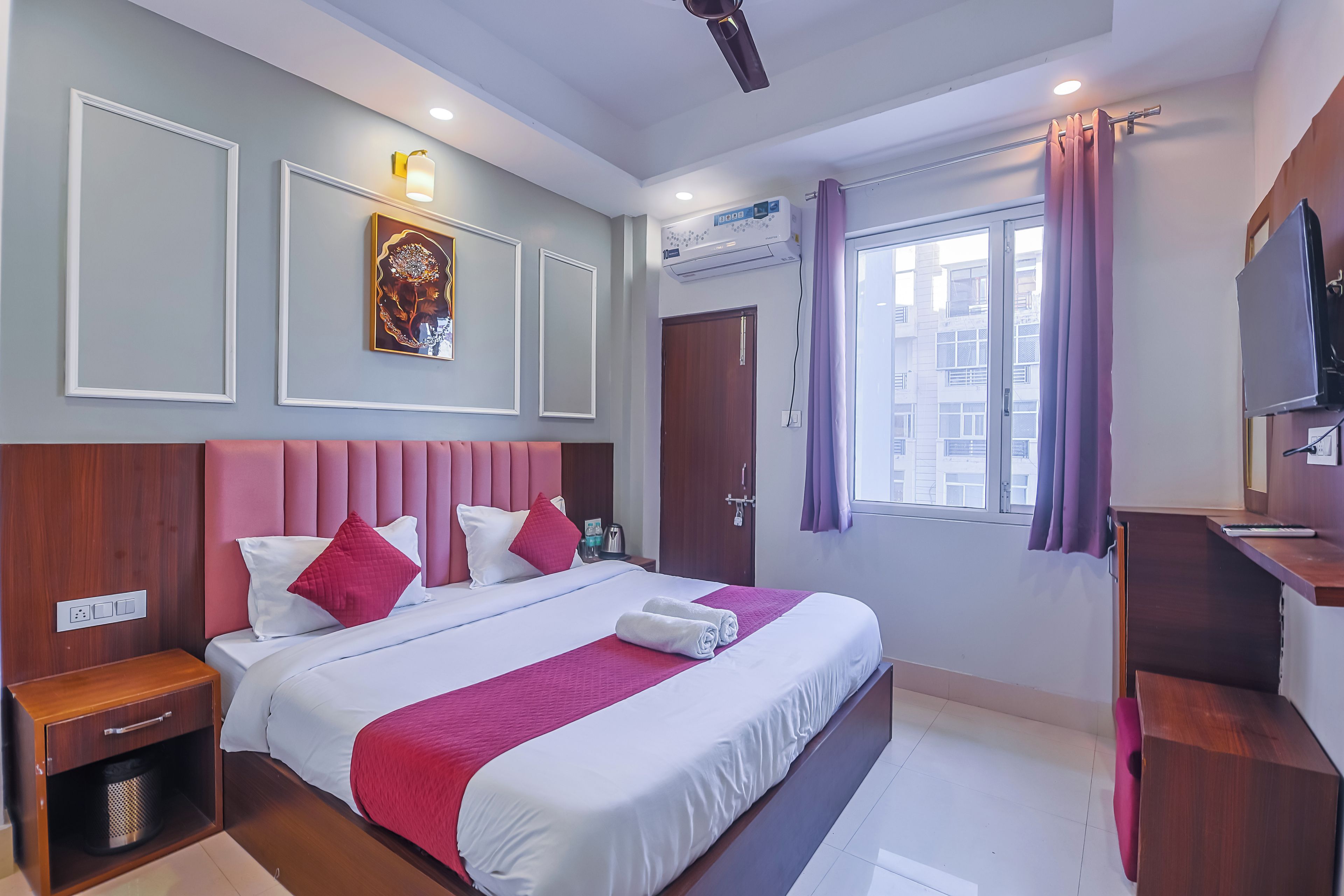 A hotel room with a large bed, a wooden nightstand, a built-in TV, and a window with curtains.