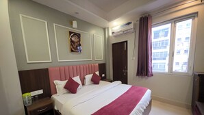 A hotel room with a large bed, a nightstand, a wall-mounted air conditioner, and a window with curtains. - Hotel Shri Ji Palace (Haridwar)