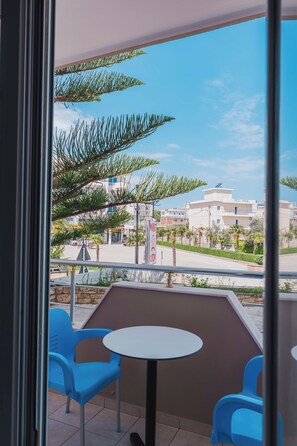 Ein Balkon mit zwei blauen Stühlen und einem kleinen Tisch, mit Blick auf eine Straße, an der sich Gebäude befinden, und einem klaren Himmel.