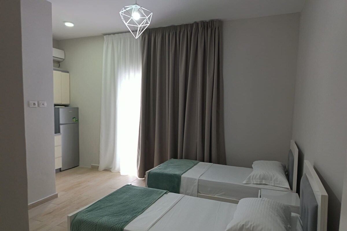 A hotel room with two beds, a kitchenette, and a large window with curtains.