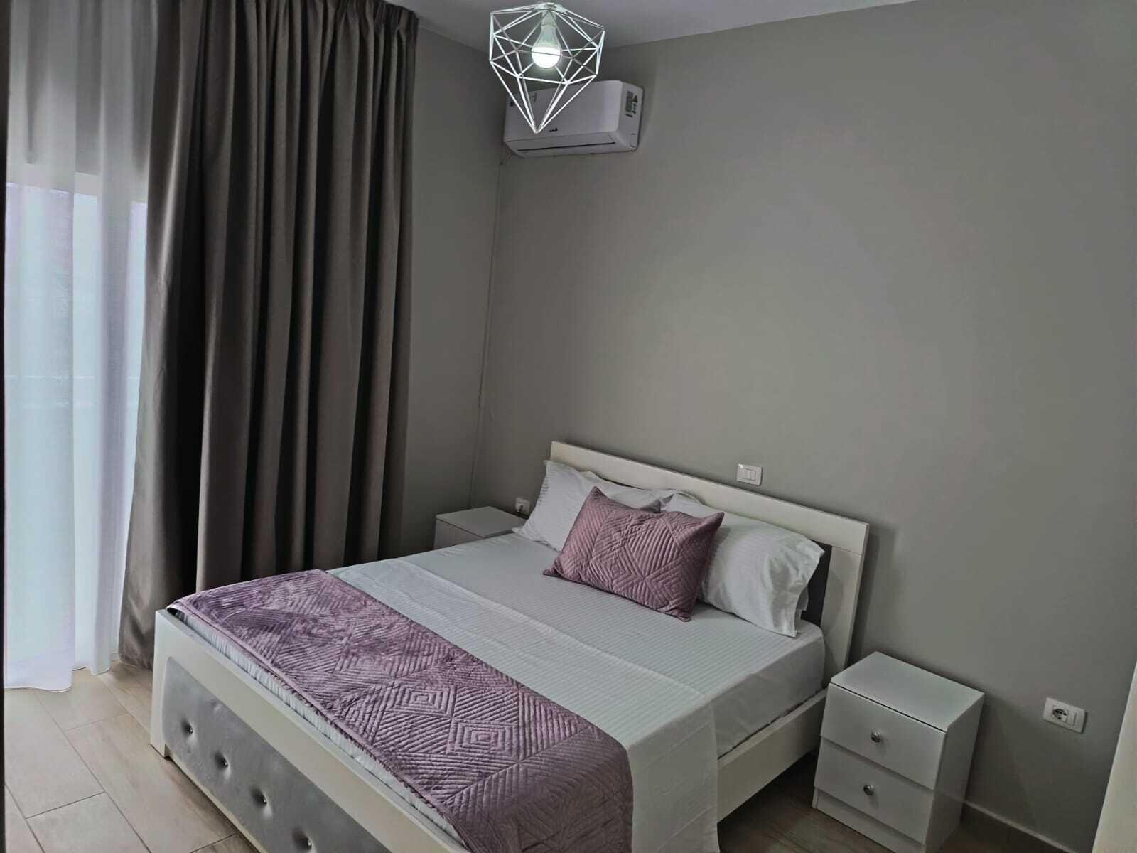 Une chambre à coucher comprenant un lit, des tables de chevet, une commode, un luminaire au design géométrique et une fenêtre avec des rideaux.