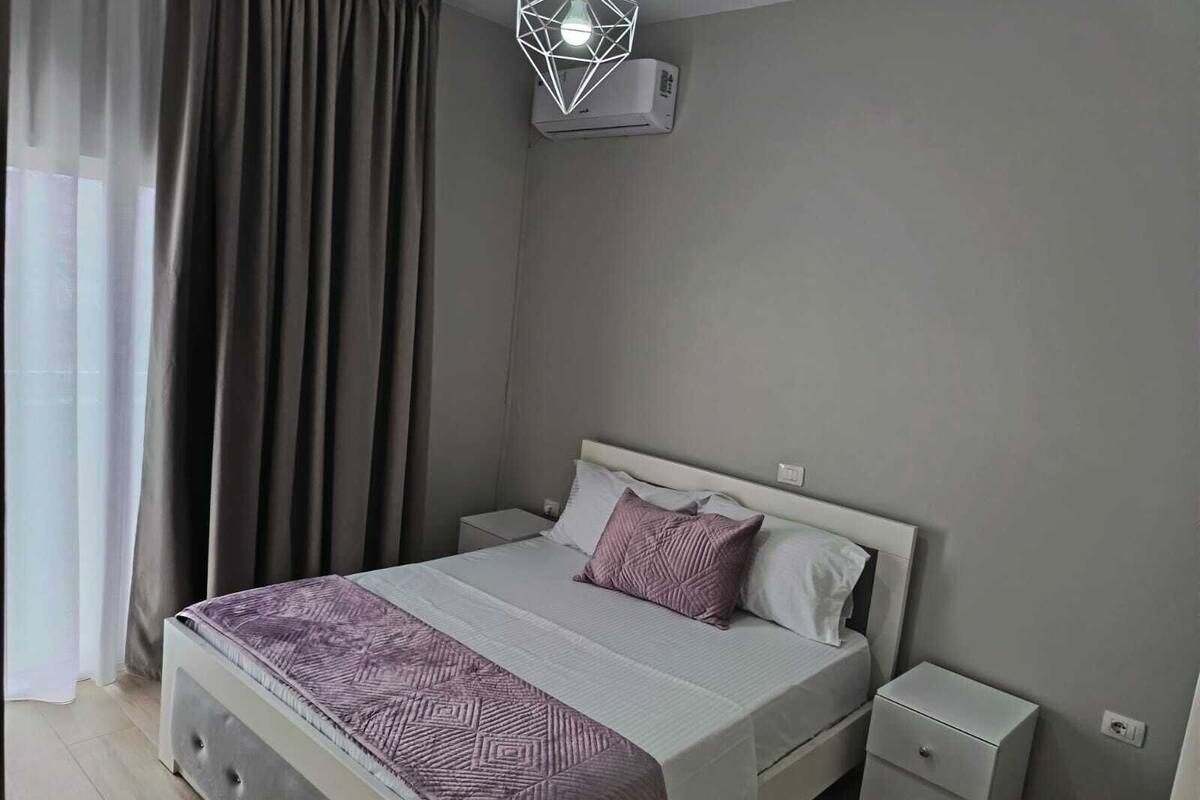 A bedroom with a bed, nightstands, a dresser, a geometric light fixture, and a window with curtains.