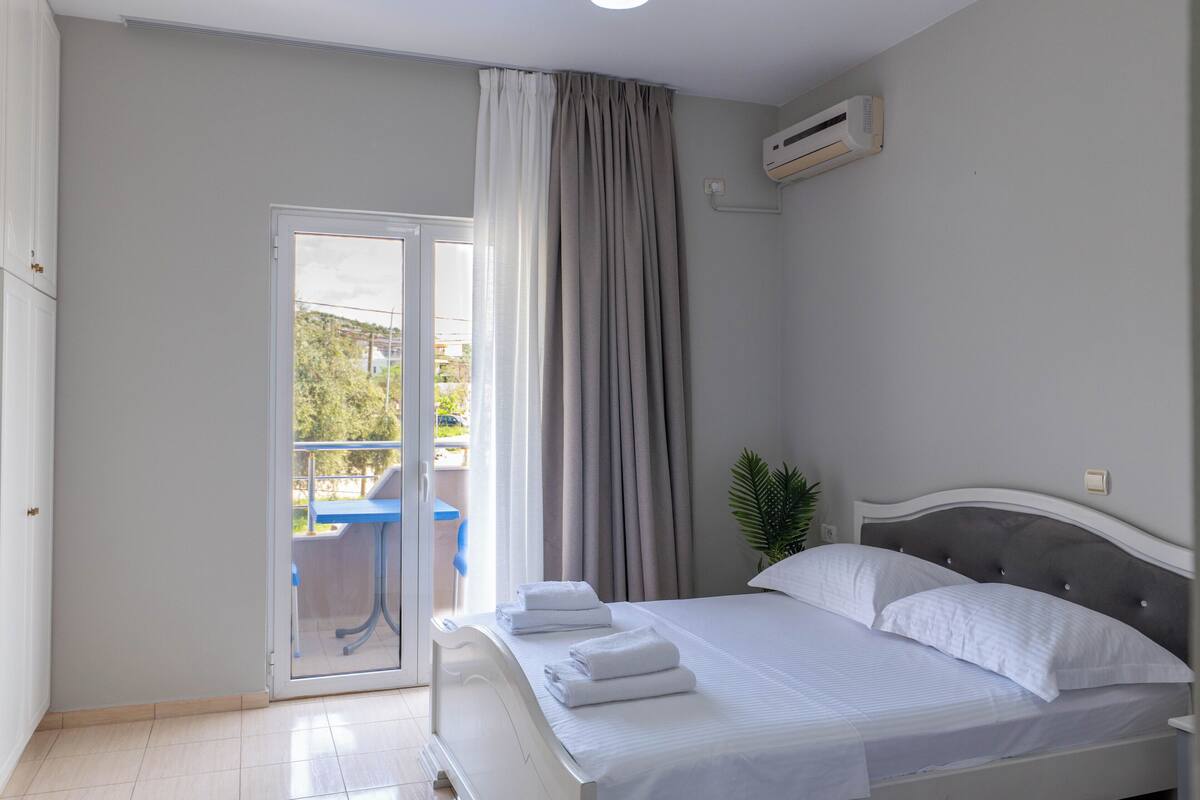 A bedroom with a bed, a window with curtains, an air conditioning unit, and a view of the outside.