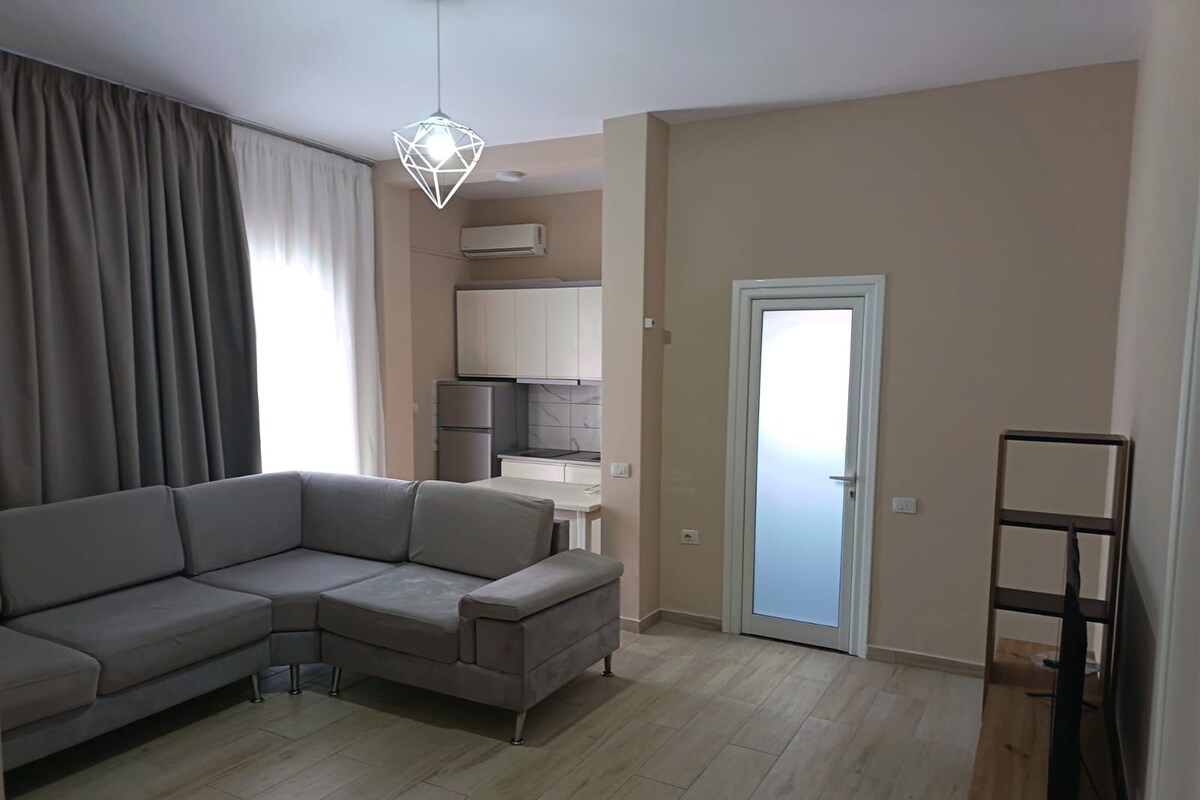 A modern living room with a grey sectional sofa, a dining table, and a kitchen area with cabinets and an air conditioner.