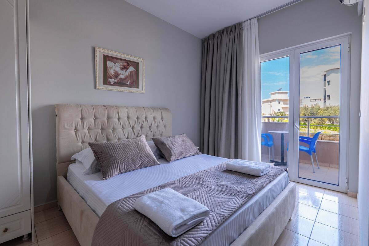 A bedroom with a large bed, a headboard, a framed picture, and a balcony with a table and chairs.
