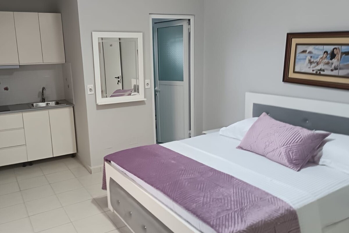 A bedroom with a bed, a nightstand, a dresser, a mirror, a door, and a kitchenette.