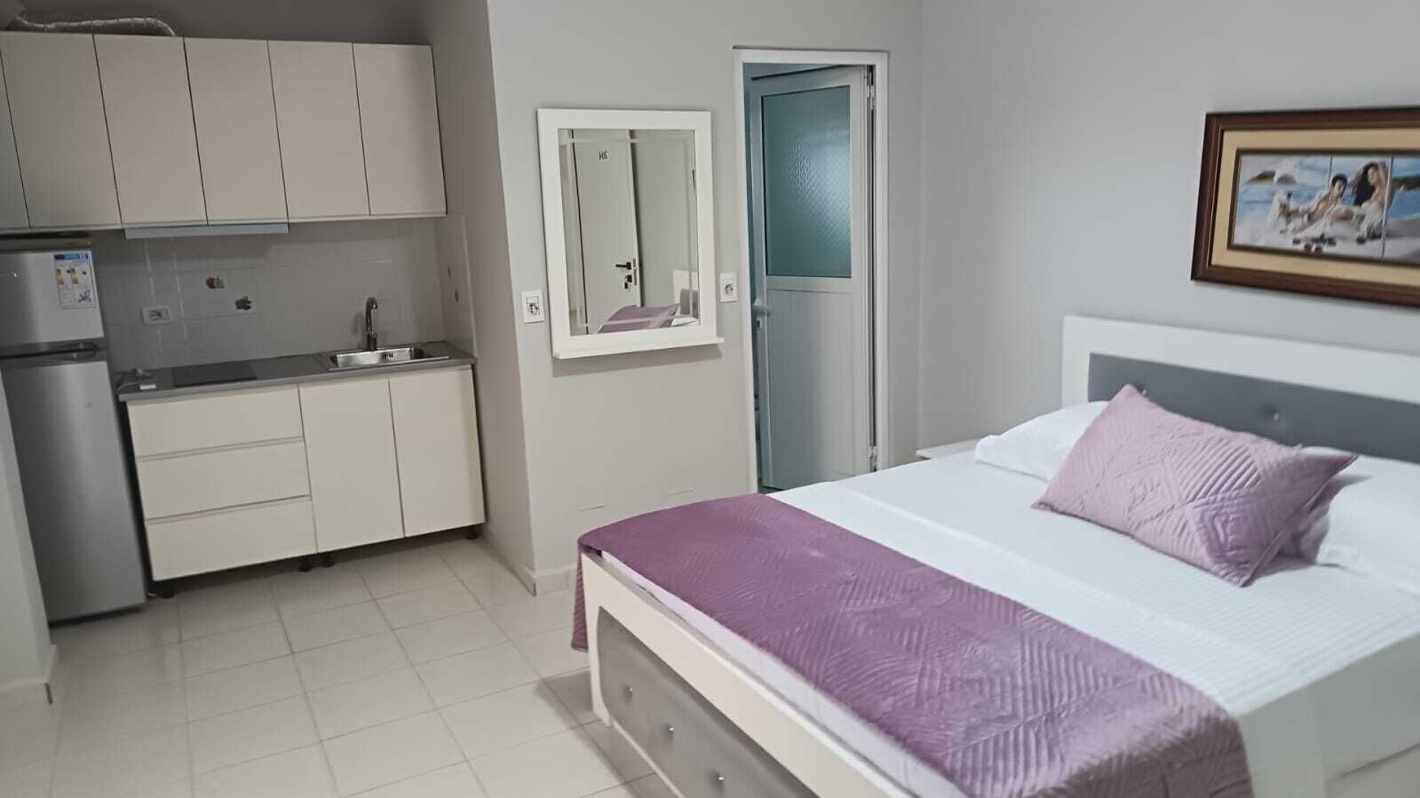 Une chambre à coucher comprenant un lit, une table de chevet, une commode, un miroir, une porte et un coin cuisine.