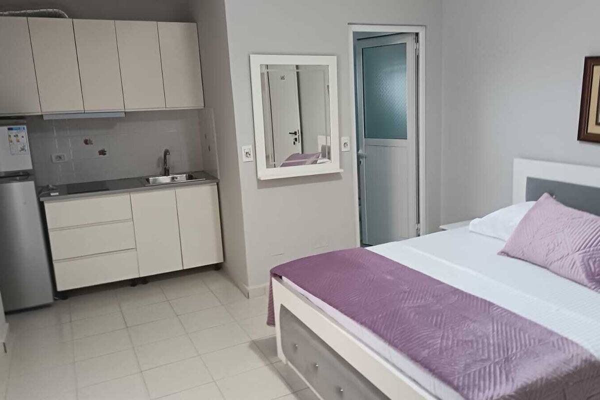 A bedroom with a bed, a nightstand, a dresser, a mirror, a door, and a kitchenette.