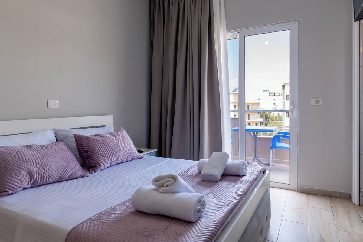 A hotel room with a bed, pillows, a window with curtains, a balcony with a chair, and a table.