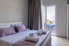 Ett hotellrum med en säng, kuddar, ett fönster med gardiner, en balkong med en stol och ett bord.