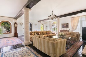 A spacious living room with a large sofa set, a coffee table, and a television. There are two large windows with curtains, a chandelier, and a clock on the wall.