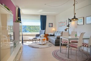 Ein modernes Wohnzimmer mit einer Couch, einem Esstisch, Stühlen, einem Glaskabinett und einem Fenster mit Vorhängen.