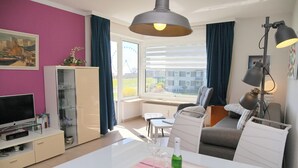A modern living room with a dining table, sofa, TV, and a large window with curtains. - Comfortable Apartment Hanseat II App. 121 with sea view and balcony in Grömitz (Grömitz)