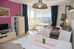 Ein modernes Wohnzimmer mit Esstisch, Sofa, Fernseher und einem großen Fenster mit Vorhängen.