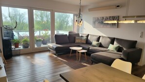 A modern living room with a sectional sofa, a dining table, and a view through large windows. - Comfortable vacation apartment Seeadler 30 with balcony near Friesenstrand in Tossens (Butjadingen-Tossens)