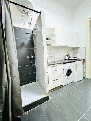 Eine kompakte Küche mit integrierter Waschmaschine und Trockner, einer Duschkabine und einer kleinen Arbeitsfläche mit Wasserkocher.