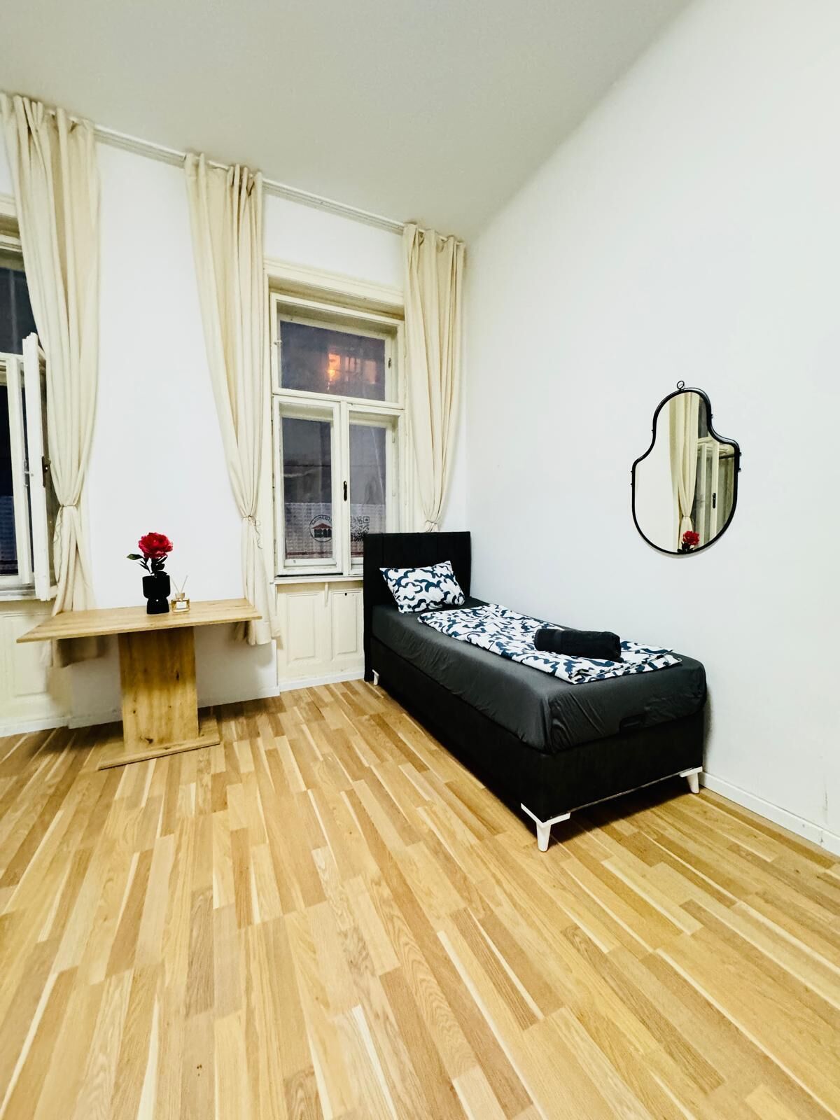 A bedroom with a bed, a bedside table, a mirror, and a window with curtains.