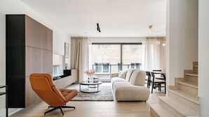 Ein modernes Wohnzimmer mit Sofa, Sessel und Couchtisch. Rechts befindet sich eine Treppe und ein großes Fenster mit Vorhängen.