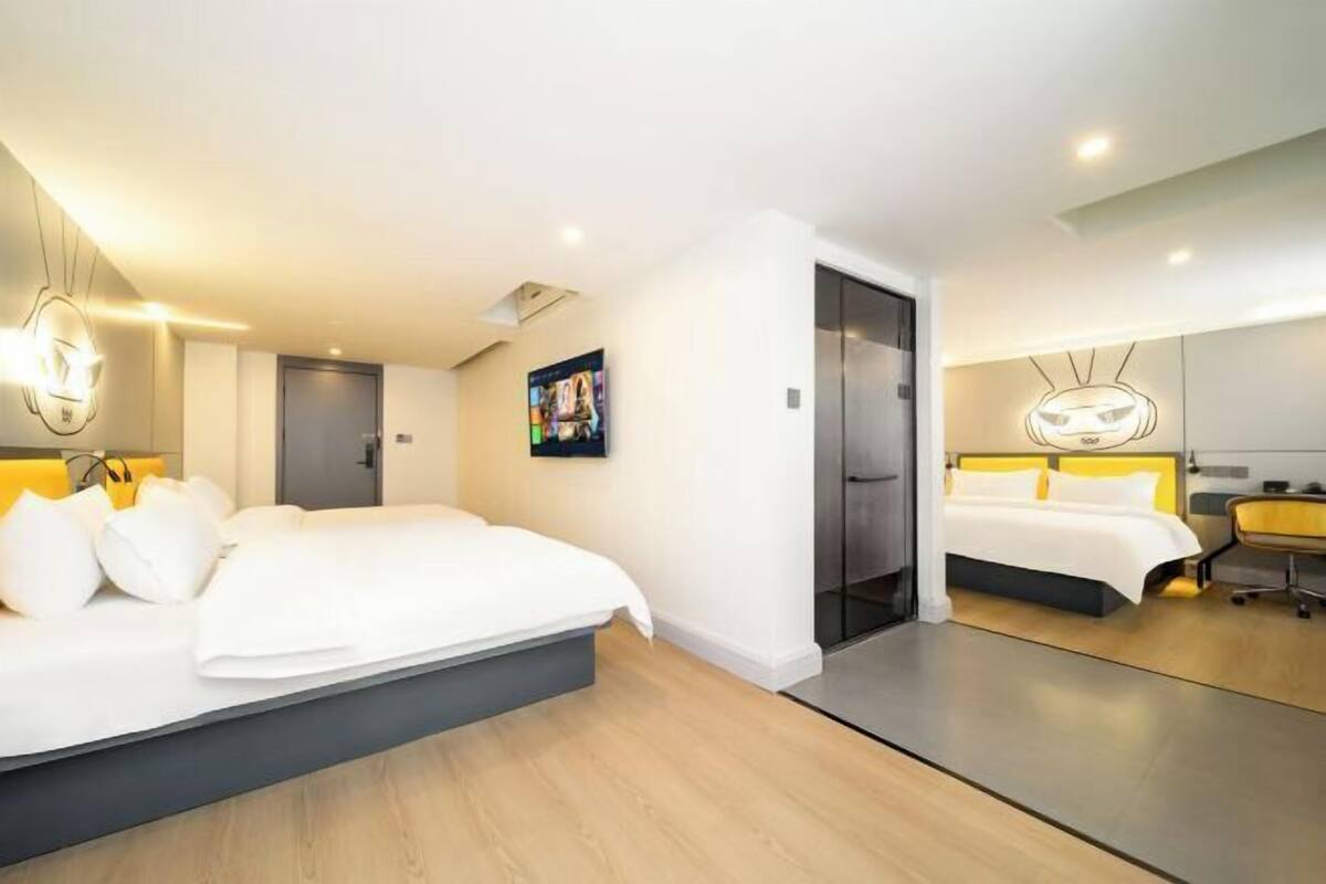 A modern hotel room with a large bed, a desk, and a chair. There is a door leading to another room and a painting on the wall.