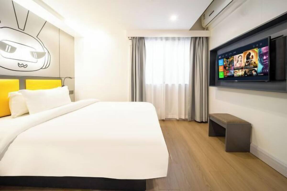 A modern hotel room with a large bed, a flat-screen TV mounted on the wall, and a bench by the window.