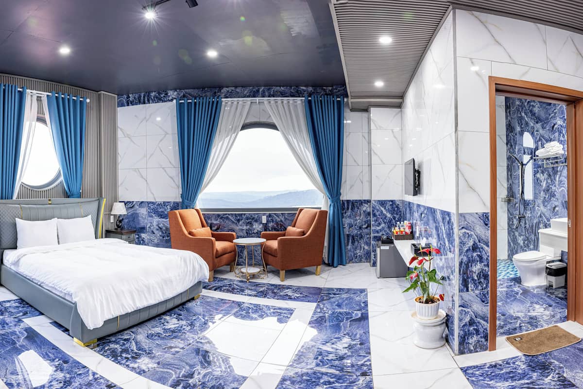 A modern hotel room with a large bed, two armchairs, a small table, a bathroom with a toilet and sink, and a view of the mountains.