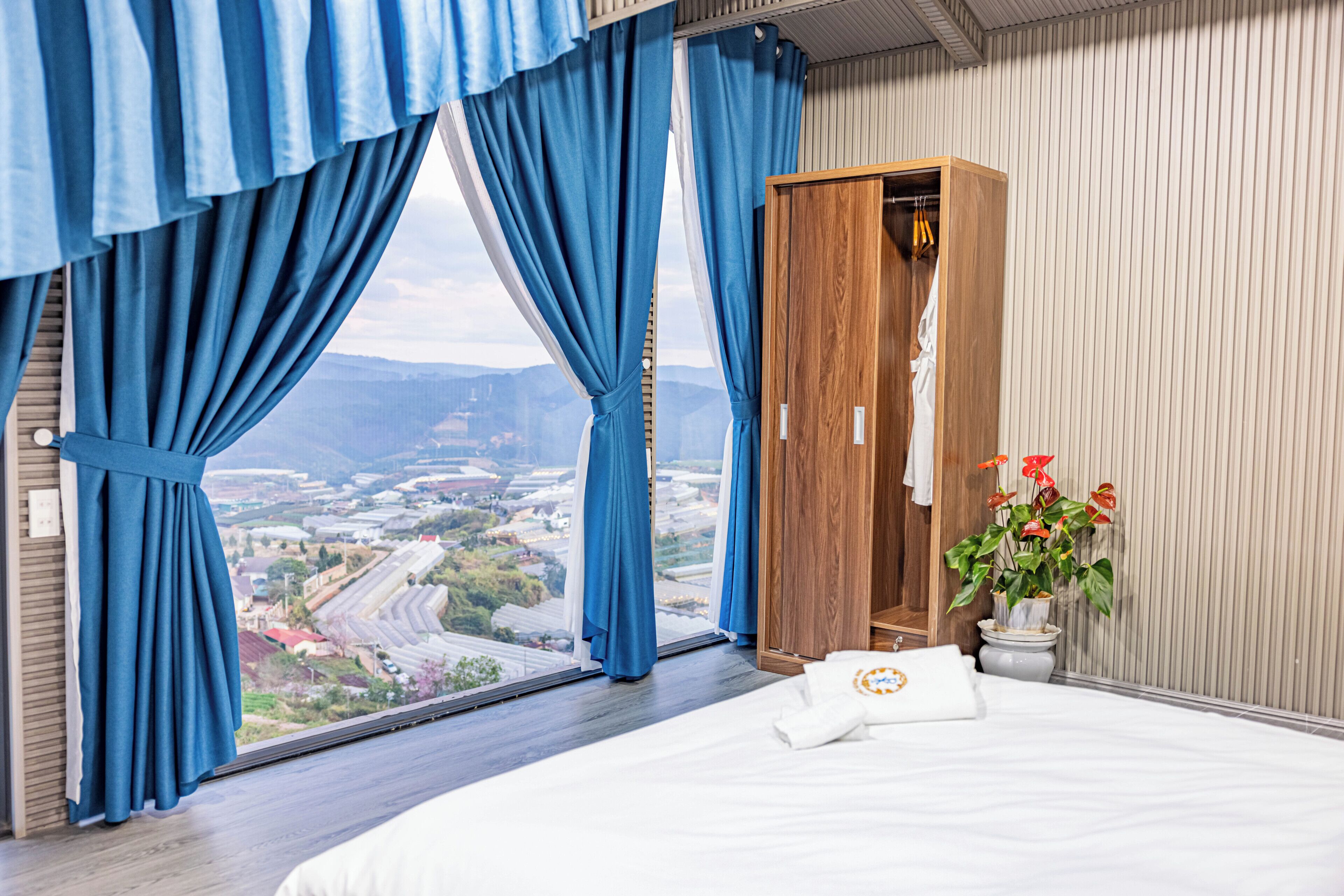 Ein Hotelzimmer mit großem Fenster, einem Holzschrank, einem Bett mit weißem Kopfkissen und einem Blumentopf mit roten Blüten.