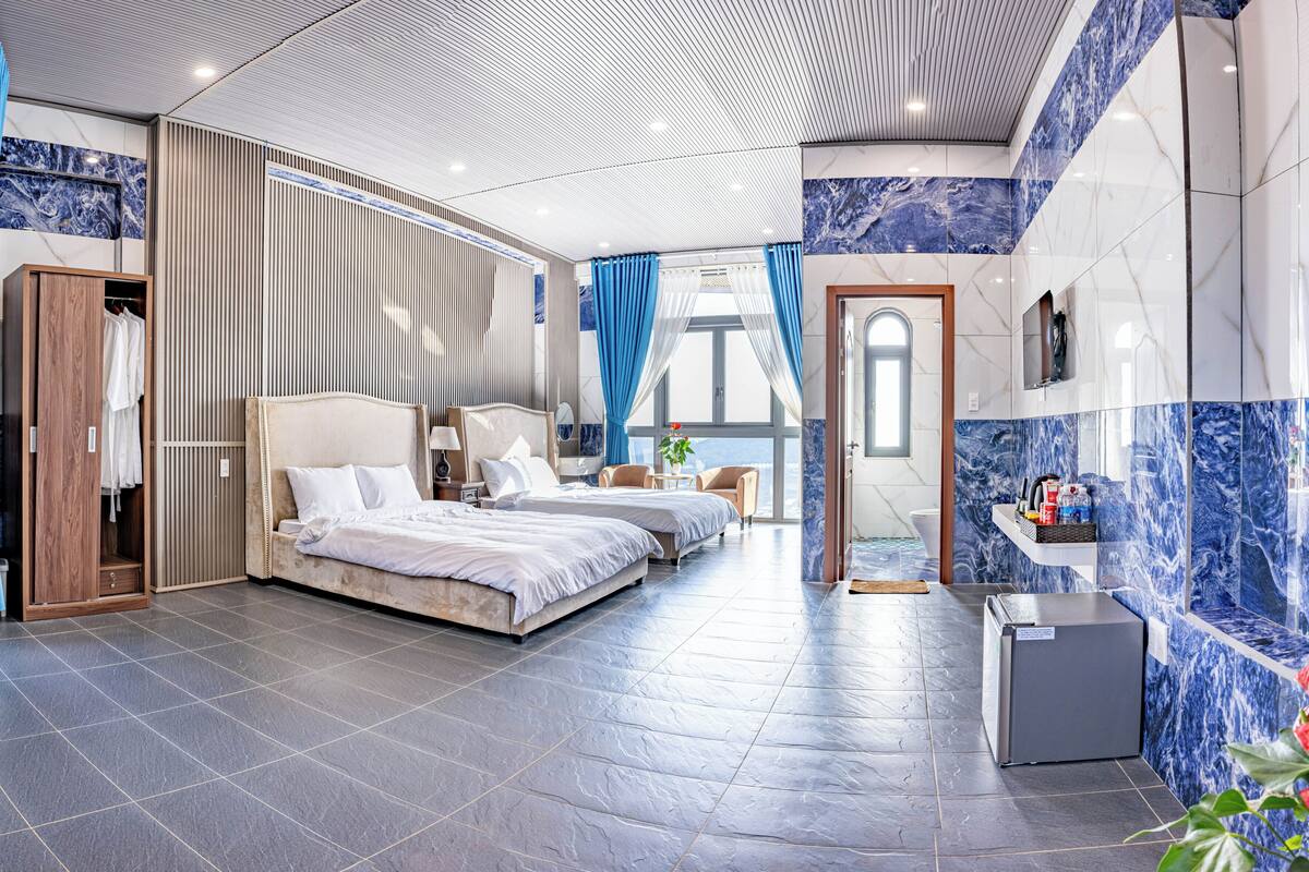 A modern hotel room with a large bed, a separate seating area, a bathroom with a marble wall, and a view of the sea.