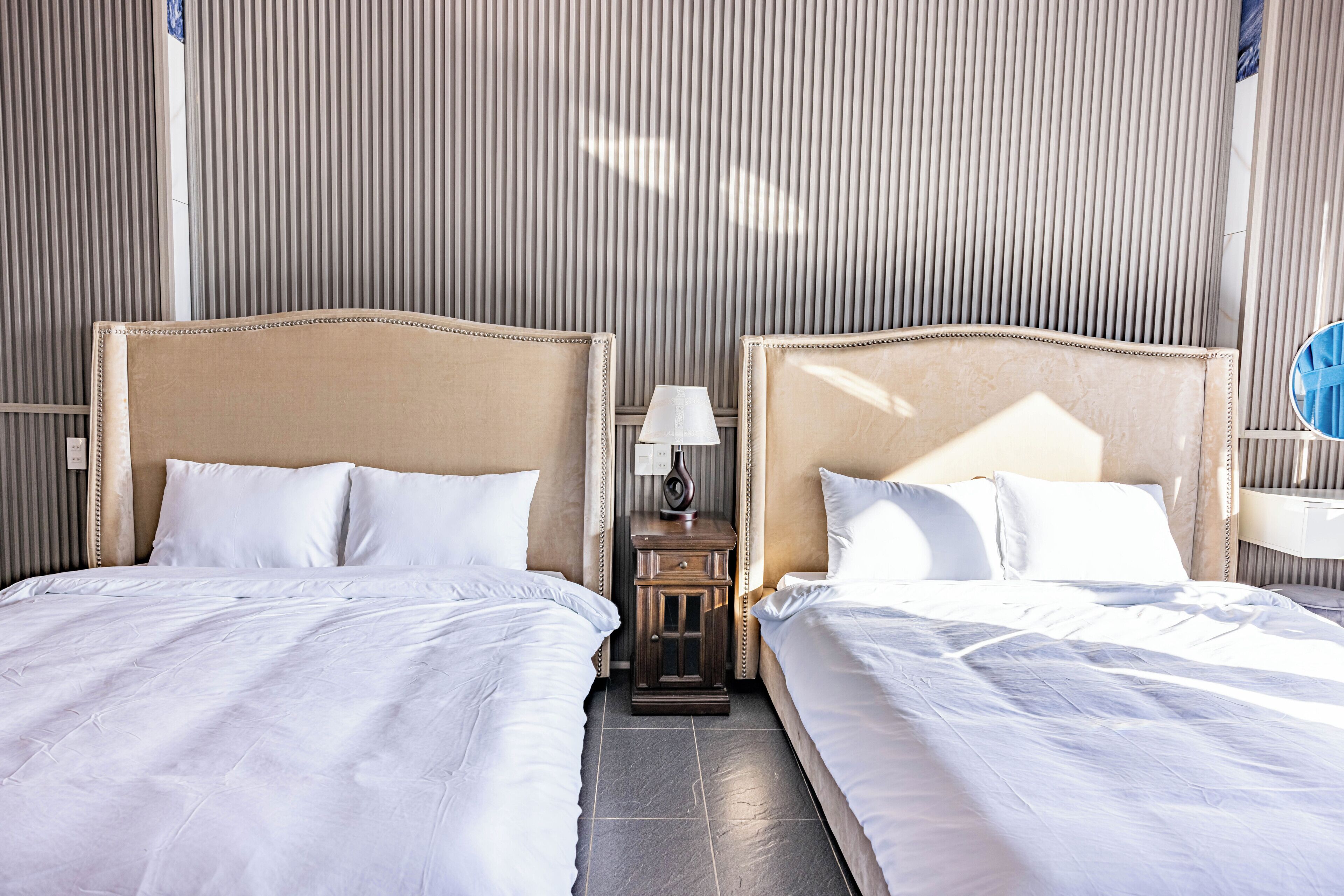 A hotel room with two beds, a nightstand with a lamp, and a window with blinds.