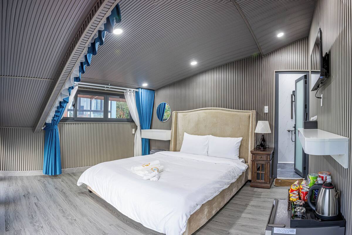 A bedroom with a bed, a bedside table, a lamp, a mirror, a shower, and a small kitchenette area.