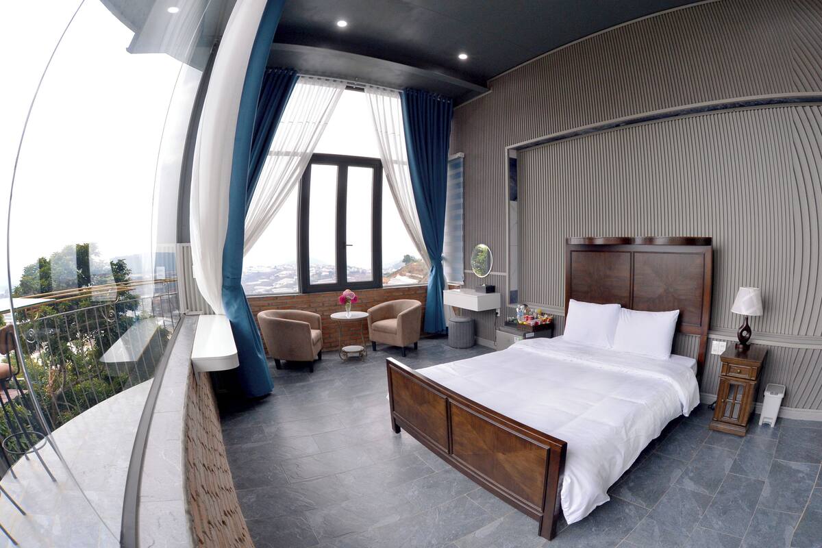 A hotel room with a large bed, a sitting area with two chairs, a small table with a vase, and a view of the city through large windows.