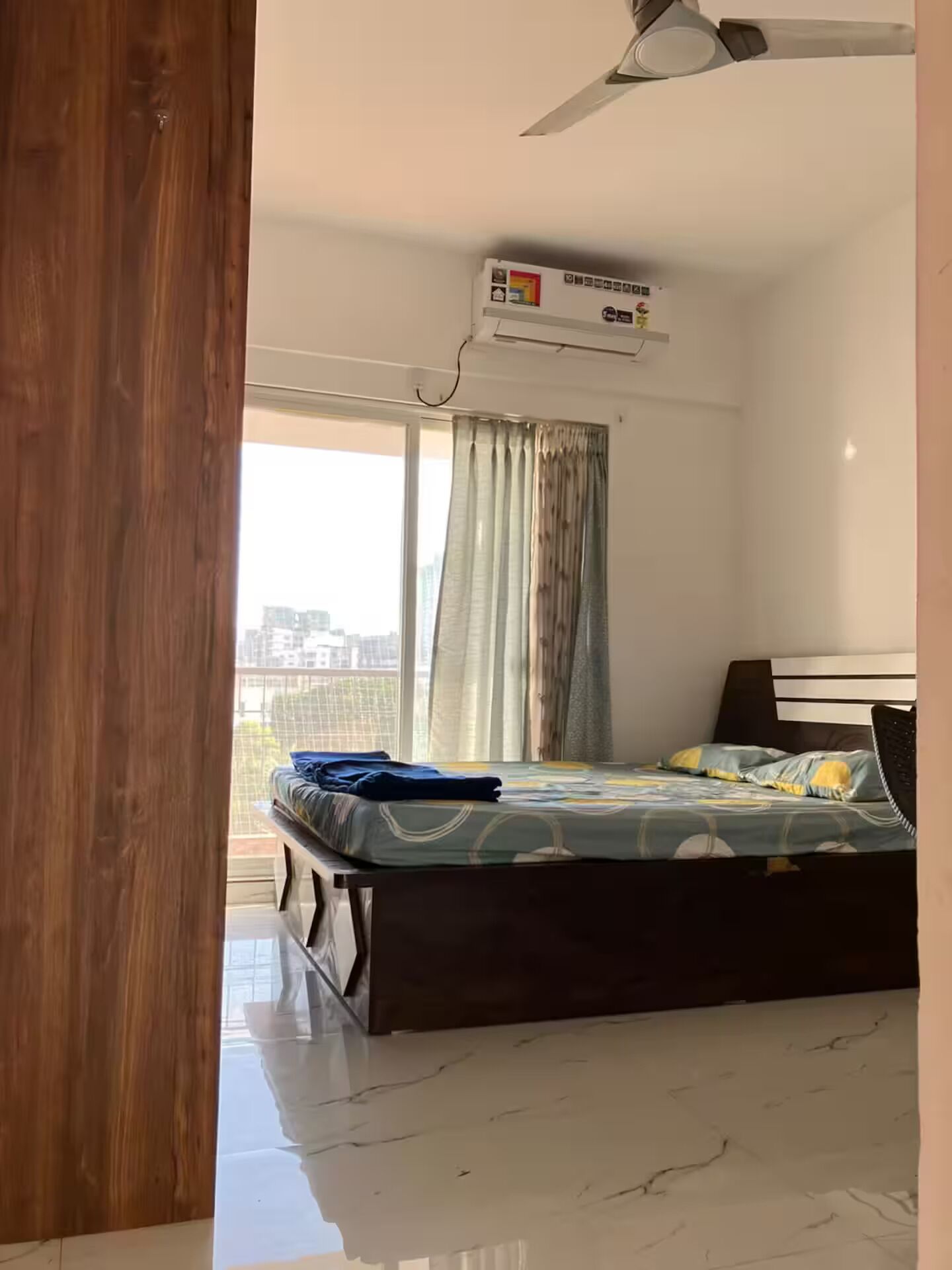 Et soveværelse med seng, aircondition, ventilator og udsigt over byen gennem store vinduer.