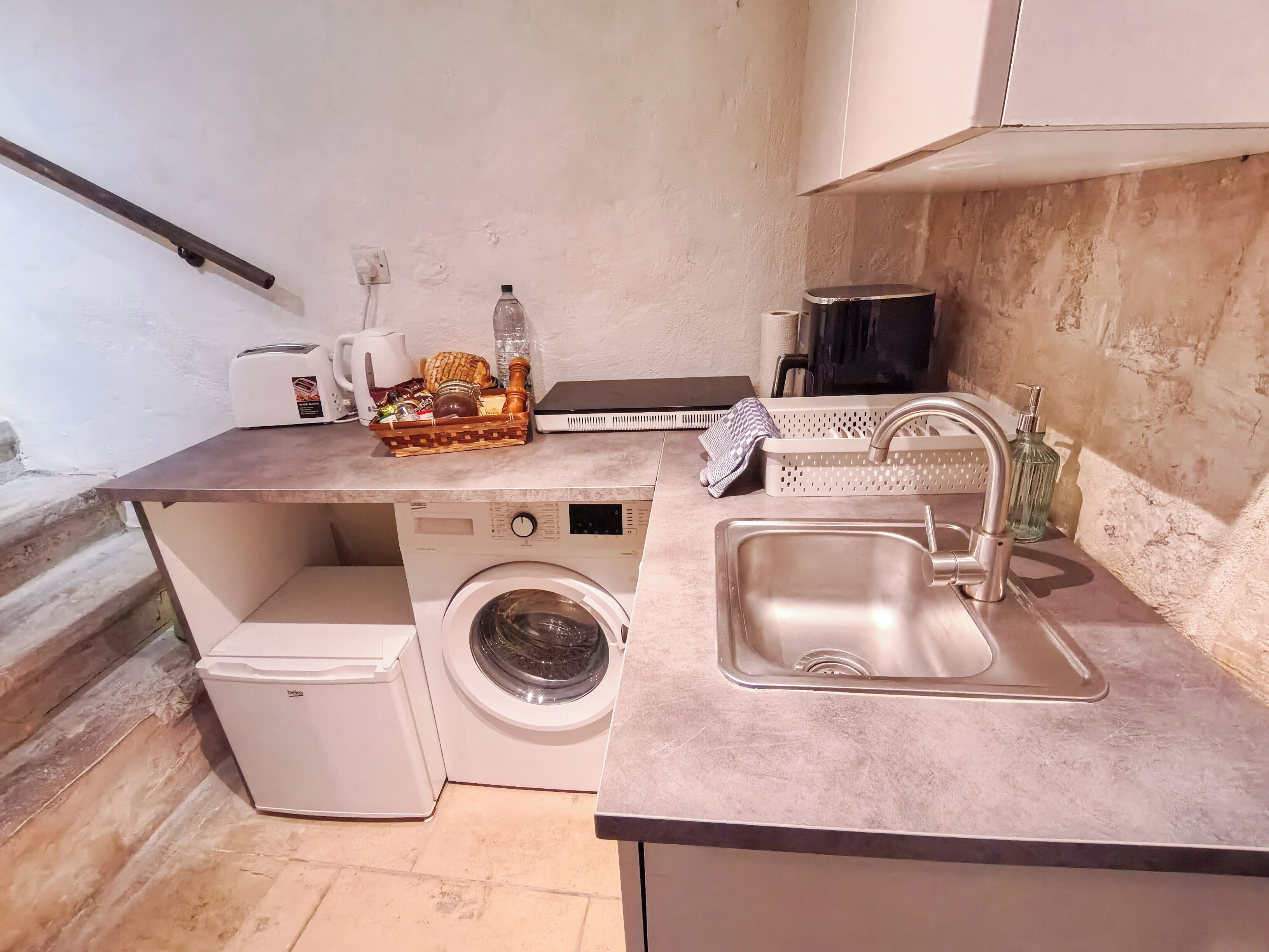 A compact kitchen with a washing machine, a sink, a toaster, and a small countertop with a basket of items.